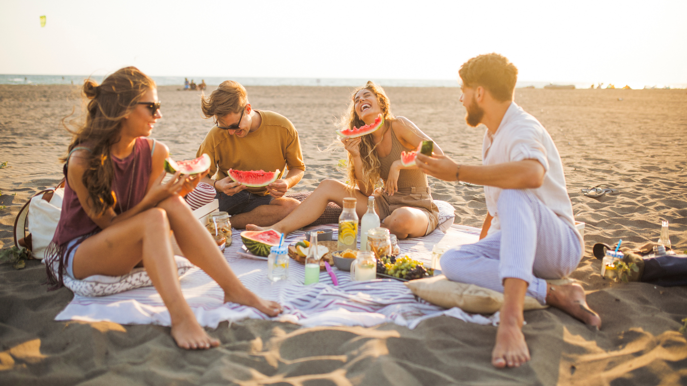 Grupa młodych ludzi robi piknik zero waste na plaży. Śmieją się i jedzą arbuza. Na kocu stoją przekąski i napoje w wielorazowych opakowaniach.